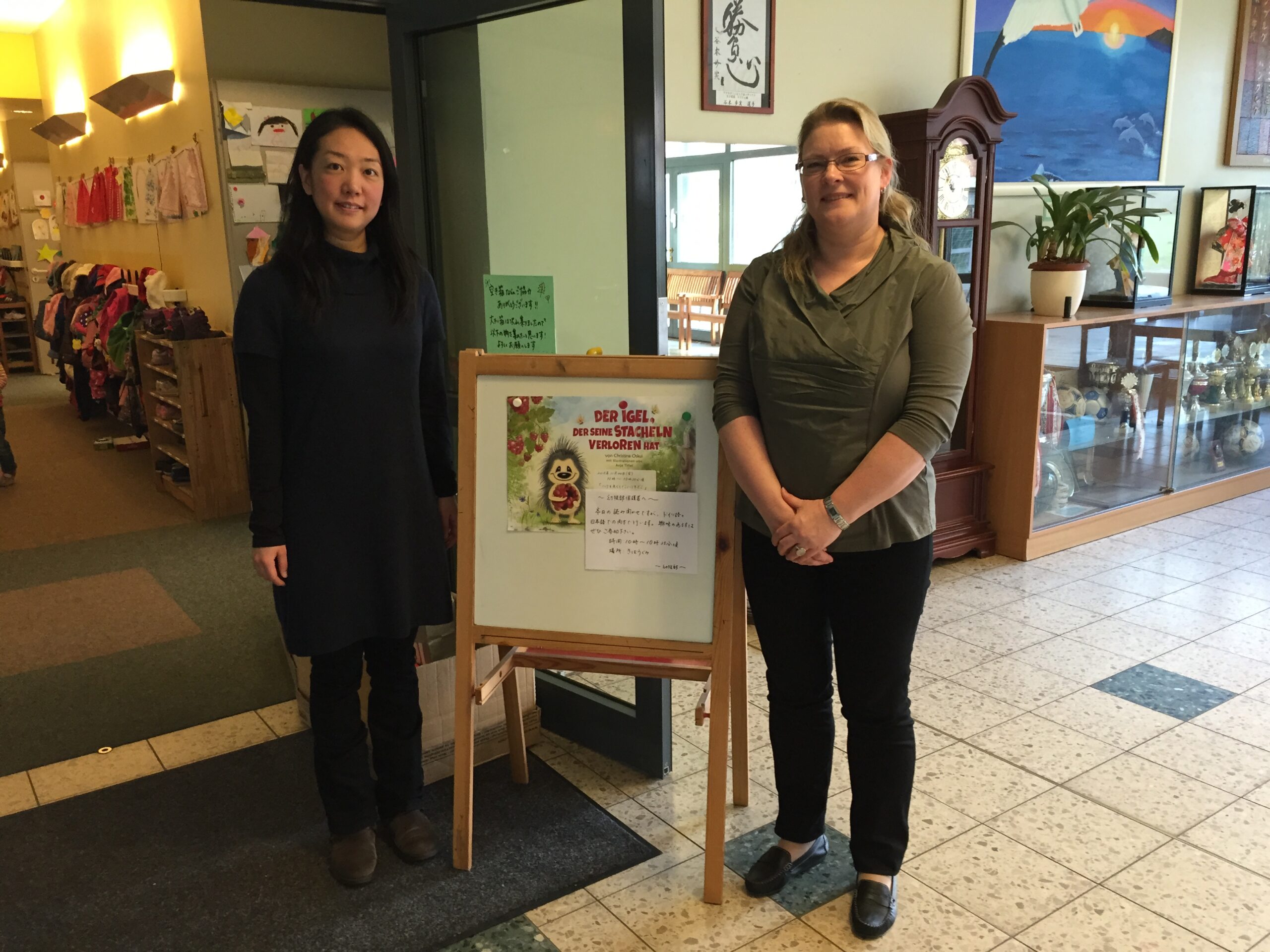 Eingangshalle japanischer Kindergarten Halstenbek. Zwei Frauen stehen neben einem Hinweisschild. Lesung Fabel "Der Igel, der seine Stacheln verloren hat!".