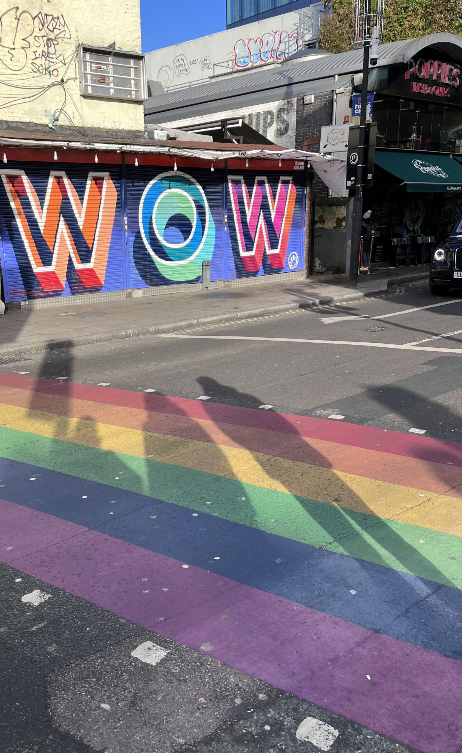 Regenbogenfarbener Zebrastreifen auf dem Schatten von Personen sind an der gegenüber liegenden Hauswand ein Grafiti mit "WOW" beschriftet.