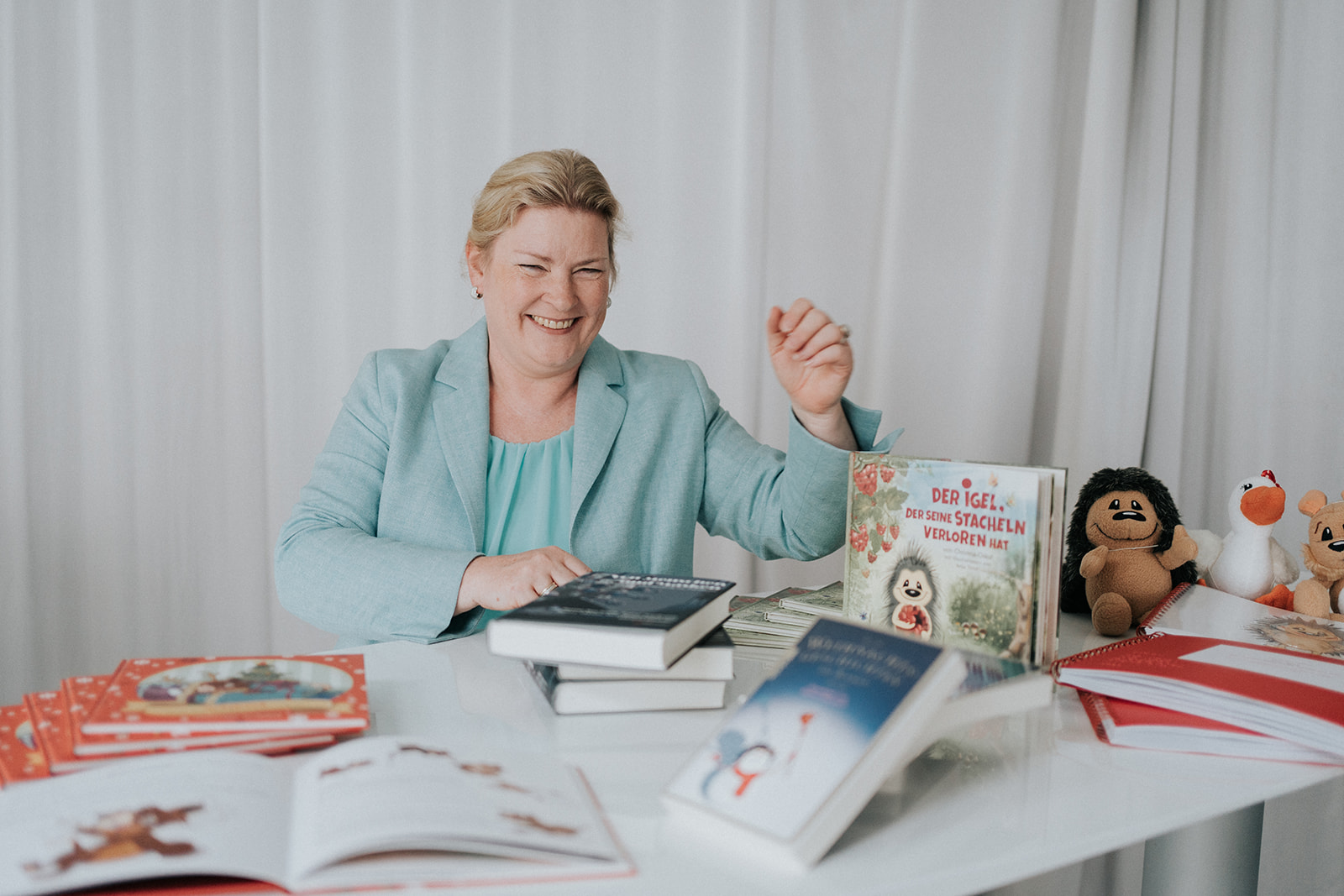 Schreibtisch mit Büchern der Autorin, Christina Oskui gefüllt, sie sitzt im Hintergrund lachend. Blonde Haare die zurückgesteckt sind, türkises Oberteil.